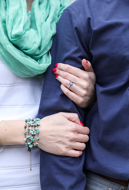Engagement photography, a closeup of a couple linking arms. She wears a turquoise scarf with a white sweater, he wears a blue shirt. 