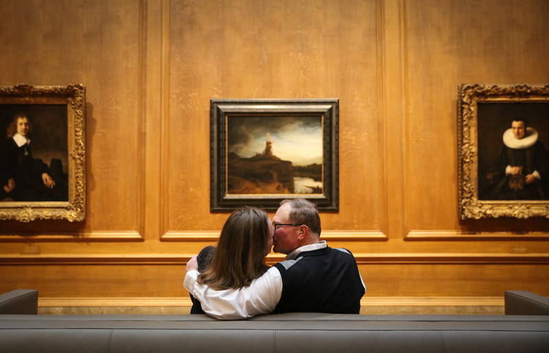 Engagement photography, a couple kisses on a couch in front of framed artwork on a wood wall. 