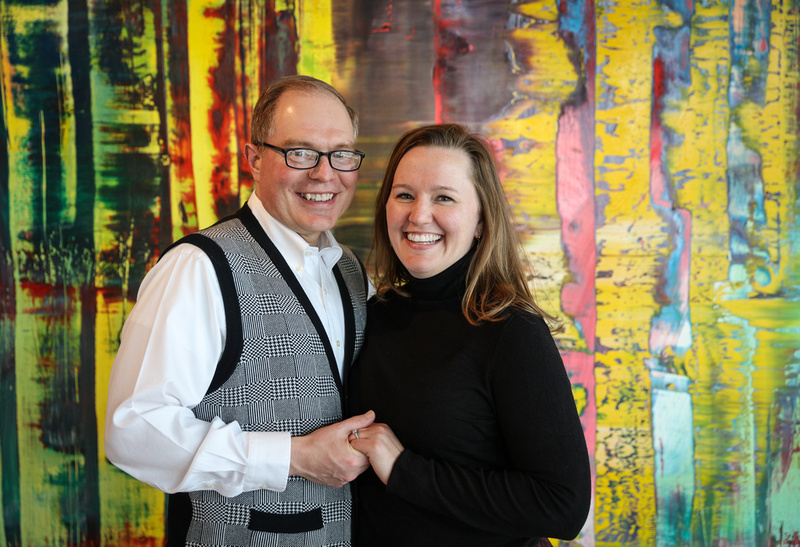 Engagement photography, a couple smiles for the camera while standing in front of a colorful piece of artwork. 