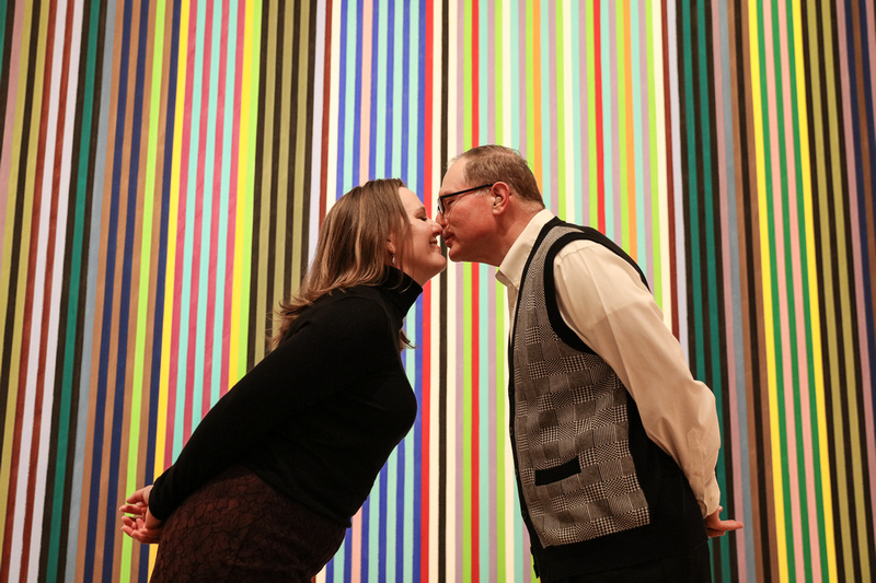 Engagement photography, a couple leans in for a kiss in front of colorful stripes on a canvas. 