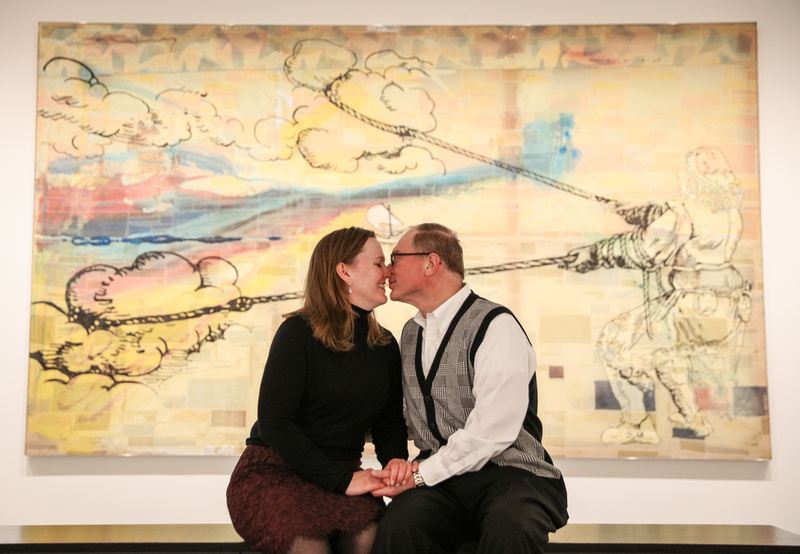 Engagement photography, a couple snuggles on a bench in front a watercolor piece of artwork. 