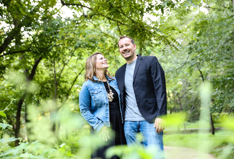 Family photography: a couple wearing jeans and black stand in the green woods, they are laughing. 