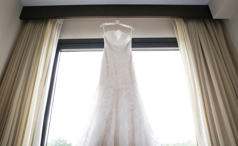 Wedding photography, a white bridal gown hangs in a picture window with beige curtains. 