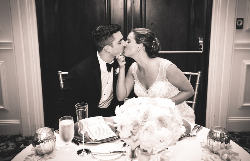 Wedding photography, a bride grabs her groom's face as they kiss sweetly at their head table. 