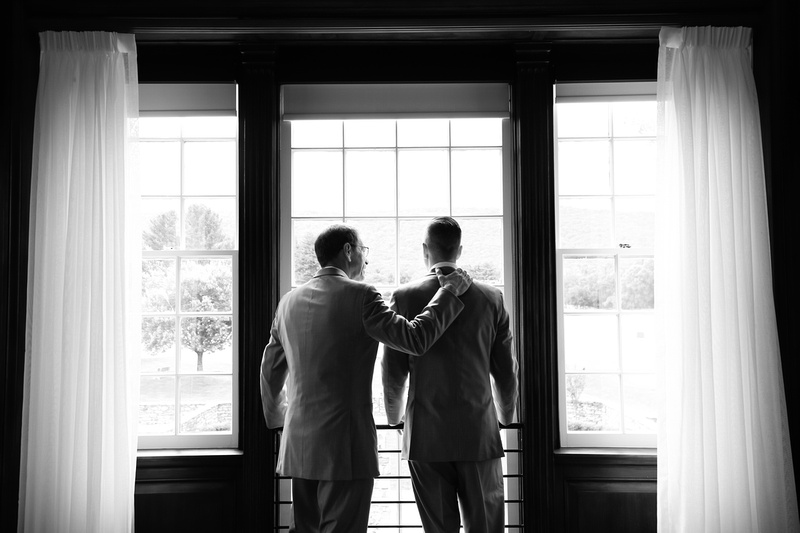 Wedding photography, a father and son lovingly stand together looking out a window at the wedding venue. 