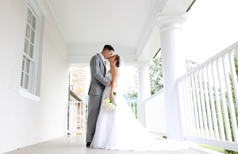 Wedding photography, a bride and groom share a romantic kiss on a white porch with trees behind them. 