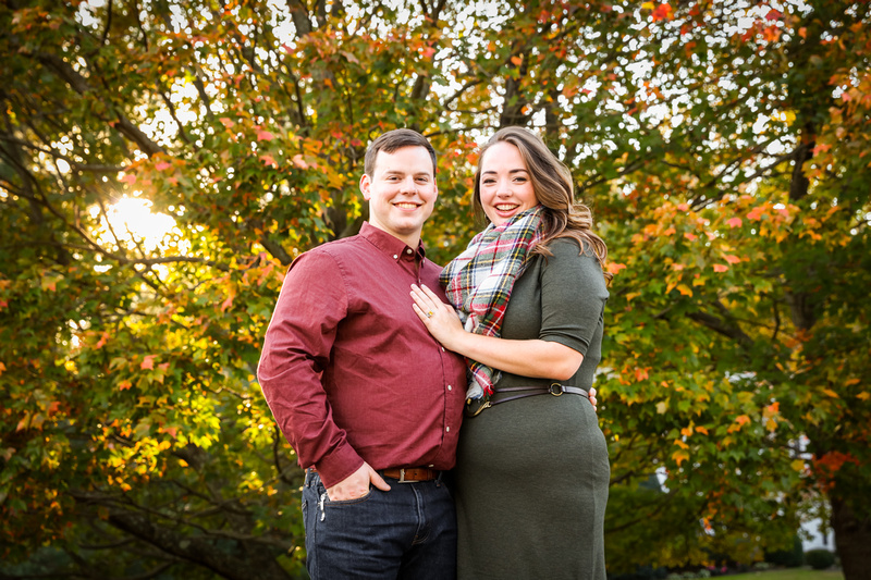 Engagement photography: a young couple smiles while standing in front of a beautiful fall tree. 