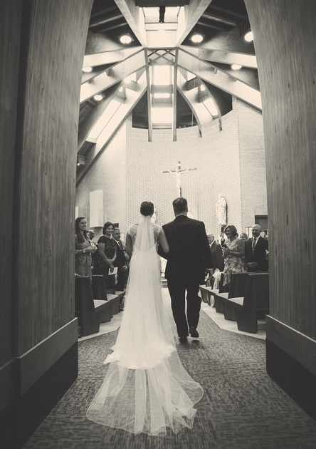 Wedding photography, a bride and her father walk down the aisle of a church. Her veil drapes down her back and trails on the floor. 