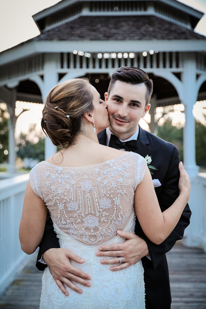 Wedding photography, a groom smiles at the camera while his bride kisses his cheek She wears an embroidered gown. 