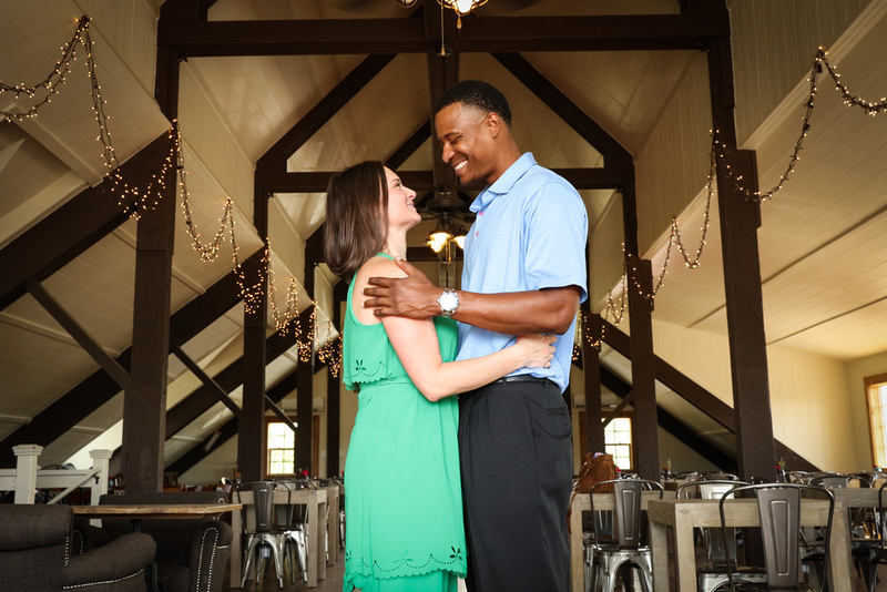 Engagement photography: a couple stands together in a barn with twinkle lights, looking at each other lovingly. 