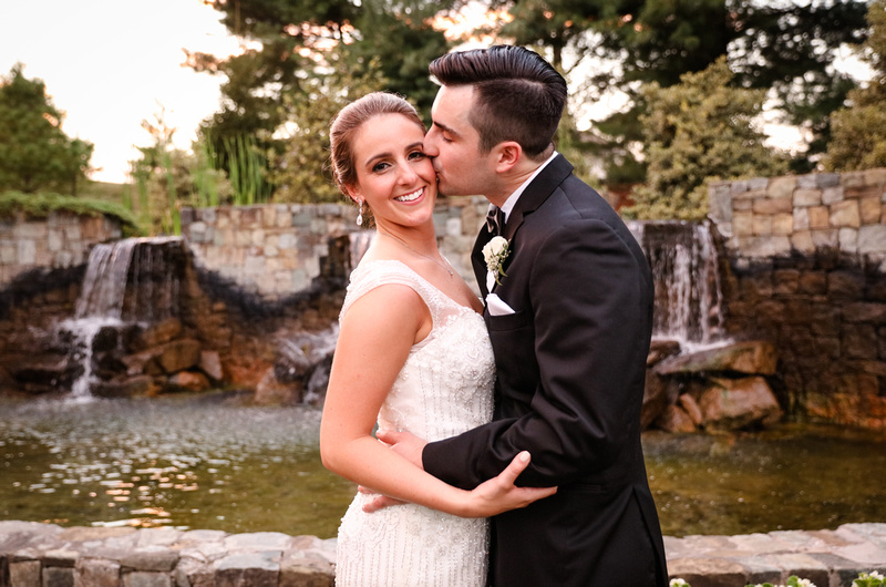 Wedding photography, a bride and groom stand in front of a large fountain. He kisses her cheek while she smiles. 