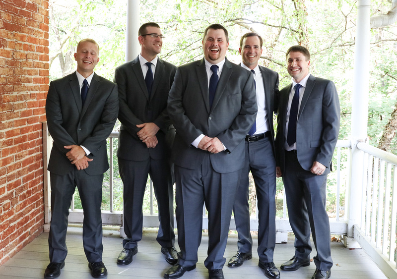Wedding photography, a groom and his groomsmen stand on a white porch with a brick wall. They are laughing and wearing grey suits. 