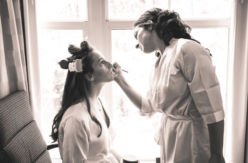 Wedding photography, two young women stand in front of a window. One wears rollers in her hair while the other does her eye makeup. 