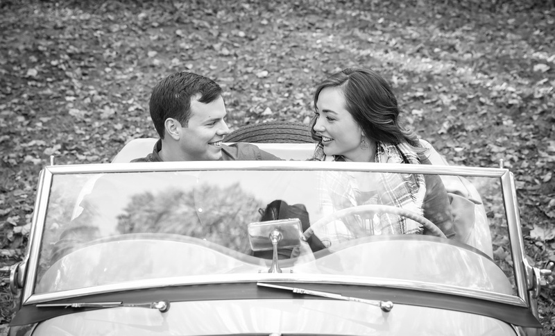 Engagement photography: a young couple sits behind the wheel of an antique car and smile at each other.