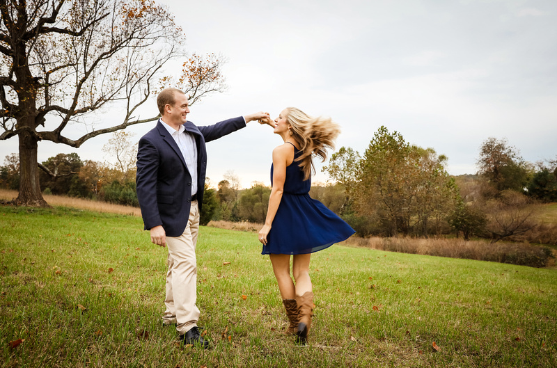 Engagement photography, a young man twirls his fiancee in a short navy dress. They are in a field. 