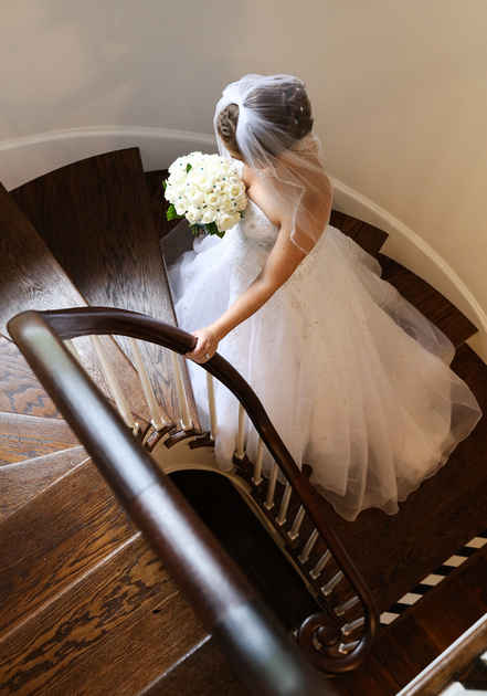 Wedding photography, a bride stands on a curved wooden stairwell with her hand on the railing, looking down at her flowers. 