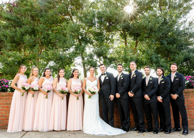 Wedding photography, a wedding party stands in front a red brick wall. They are wearing pink dresses and black tuxedos.  
