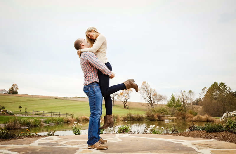 Engagement photography, a young man lifts his lady who wears boots, up into the air. She beams at him. 