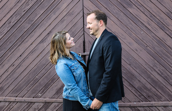Family photography: a couple stands facing each other and holding hands in front of brown barn doors. 
