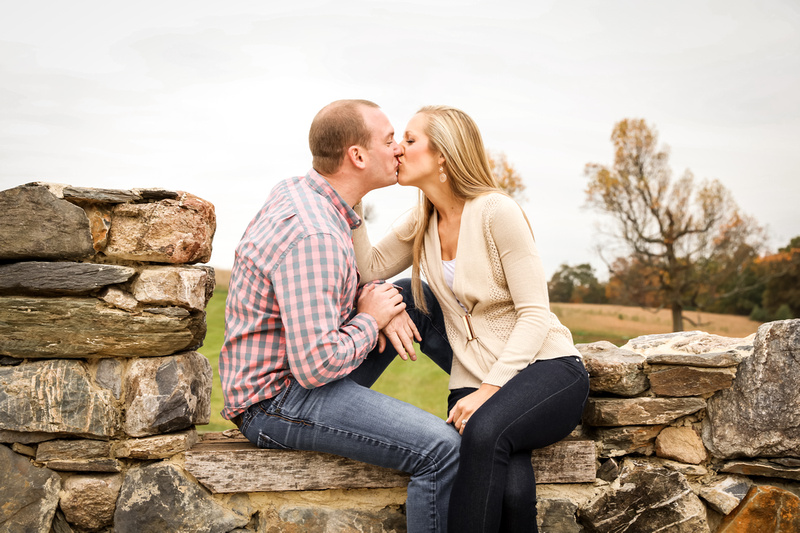 Engagement photography, a couple sneaks a kiss while sitting on a stone wall. They are wearing jeans. 