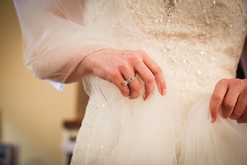 Wedding photography, a closeup of an engagement ring worn by a bride in a white gown with beaded embellishment. 
