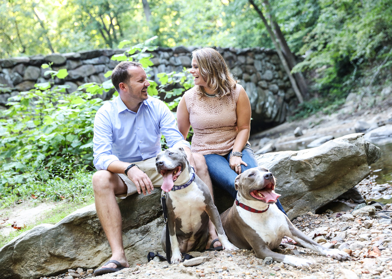Family photography: a man and woman laugh as they sit in the woods with their two pit bulls. 