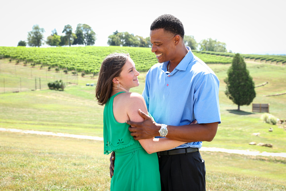 Engagement photography: a couple stand in front of an overlook of the vineyard, she glances up at her fiancé lovingly as he embraces her. 
