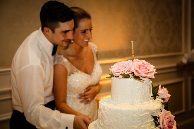 Wedding photography, the bride and groom smile while cutting their wedding cake. It is covered with pink roses. 