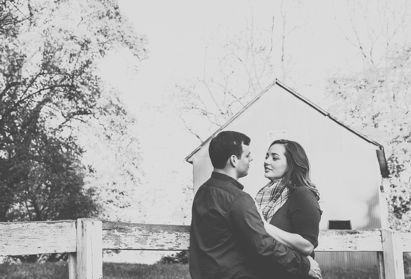 Engagement photography: a couple stands in front of a split rail fence and a barn. 