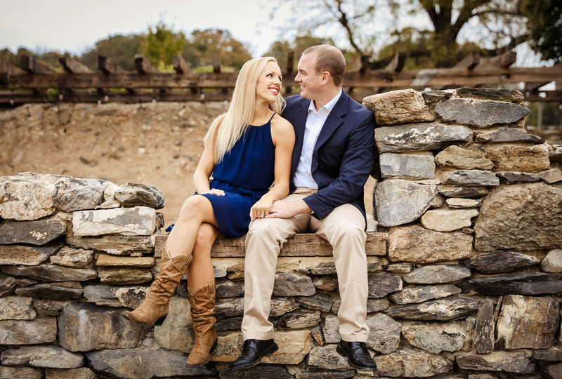 Engagement photography, a couple sits on stone wall with trees behind them. 