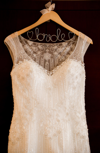 Wedding photography, a white embroidered gown hangs on a wooden hanger that says bride. 