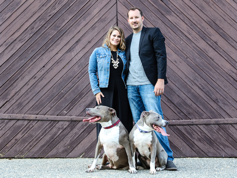 Family photography: a couple stands in front of brown barn doors with the pit bulls sitting at their feet. 