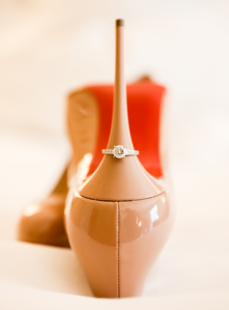 Wedding photography, a pair of nude high heels with a red bottom on a bed. On the heel is a diamond engagement ring. 