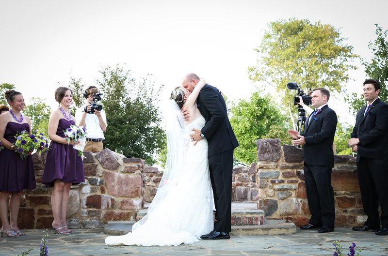 Wedding photography, a bride and groom share their first kiss as the wedding party smiles on. 