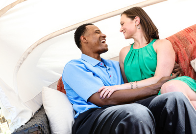 Engagement photography: a man in a blue shirt laughs adoringly at his fiancé, they sit on a red couch. 