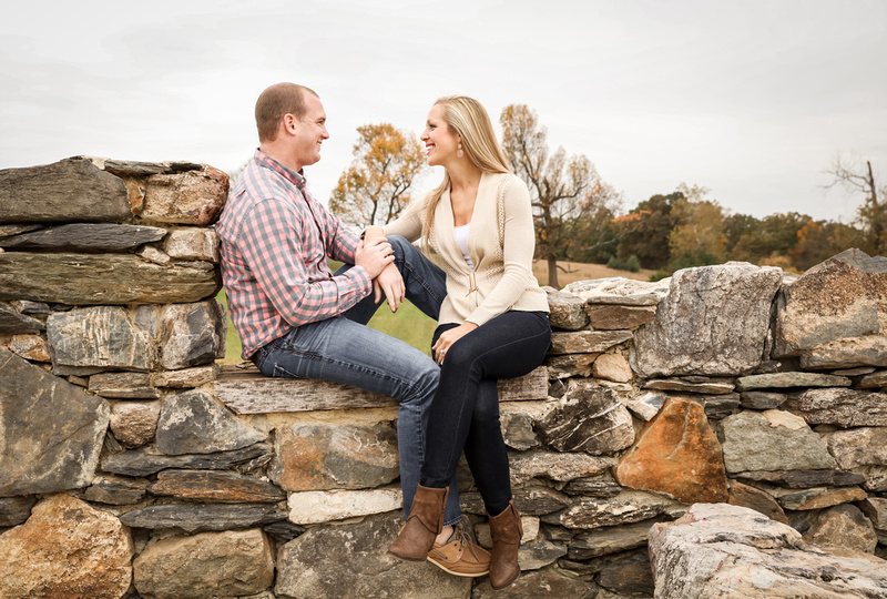 Engagement photography, two young loves sit on a stone wall smiling at each other. 