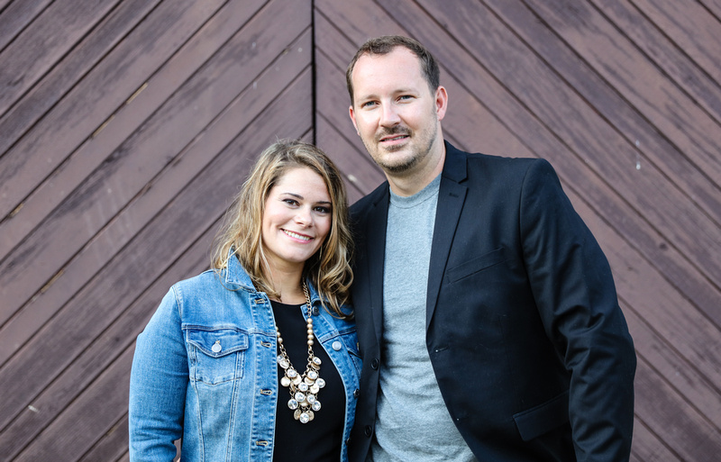 Family photography: a couple stands in front of brown barn doors. She wears a jean jacket with a gold necklace. We wears a black blazer. 