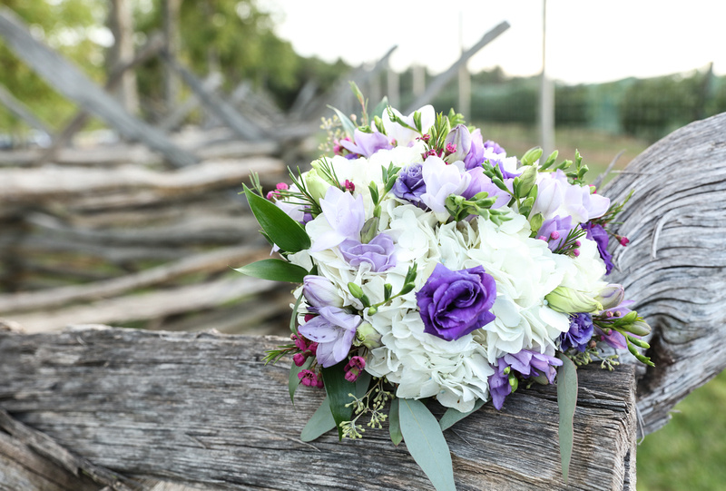Wedding photography, a bouquet of white, purple, green, and magenta flowers sits on a wooden fence. 