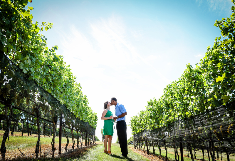 Engagement photography: a couple lean in for a kiss, while holding wine glasses in a long corridor of grapes at a Virginia vineyard.  