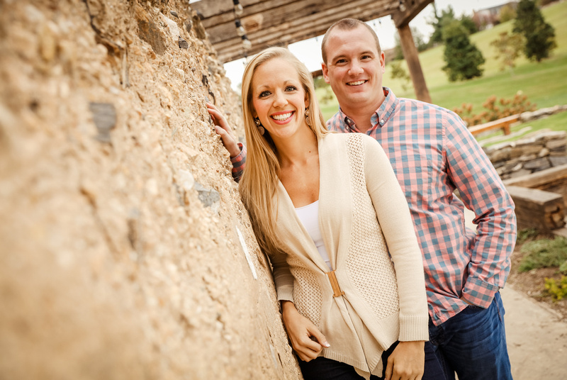 Engagement photography, a couple leans against a stone wall. She wears a cream sweater, he wears a plaid shirt. 