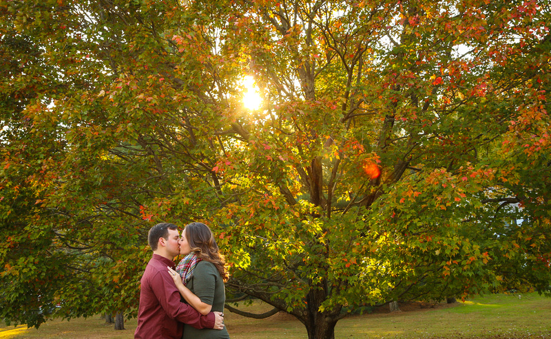 Engagement photography: a young couple kisses while standing in front of a beautiful fall tree. 